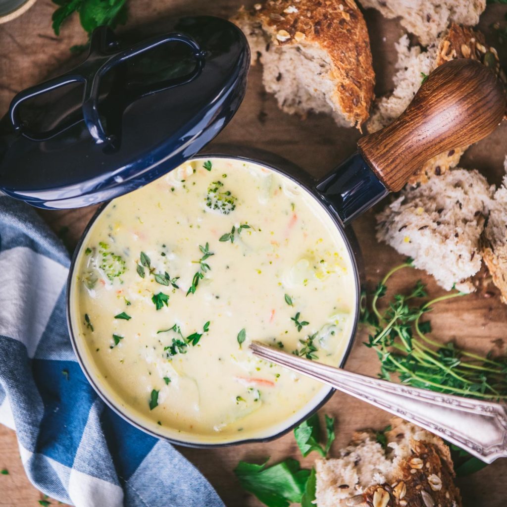 Square overhead shot of a bowl of broccoli and cheese soup on a wooden board with bread