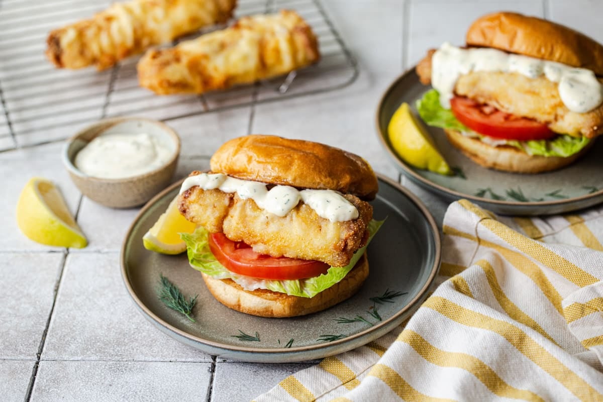 Horizontal side shot of a crispy fried fish sandwich on a plate with homemade tartar sauce and a lemon wedge.