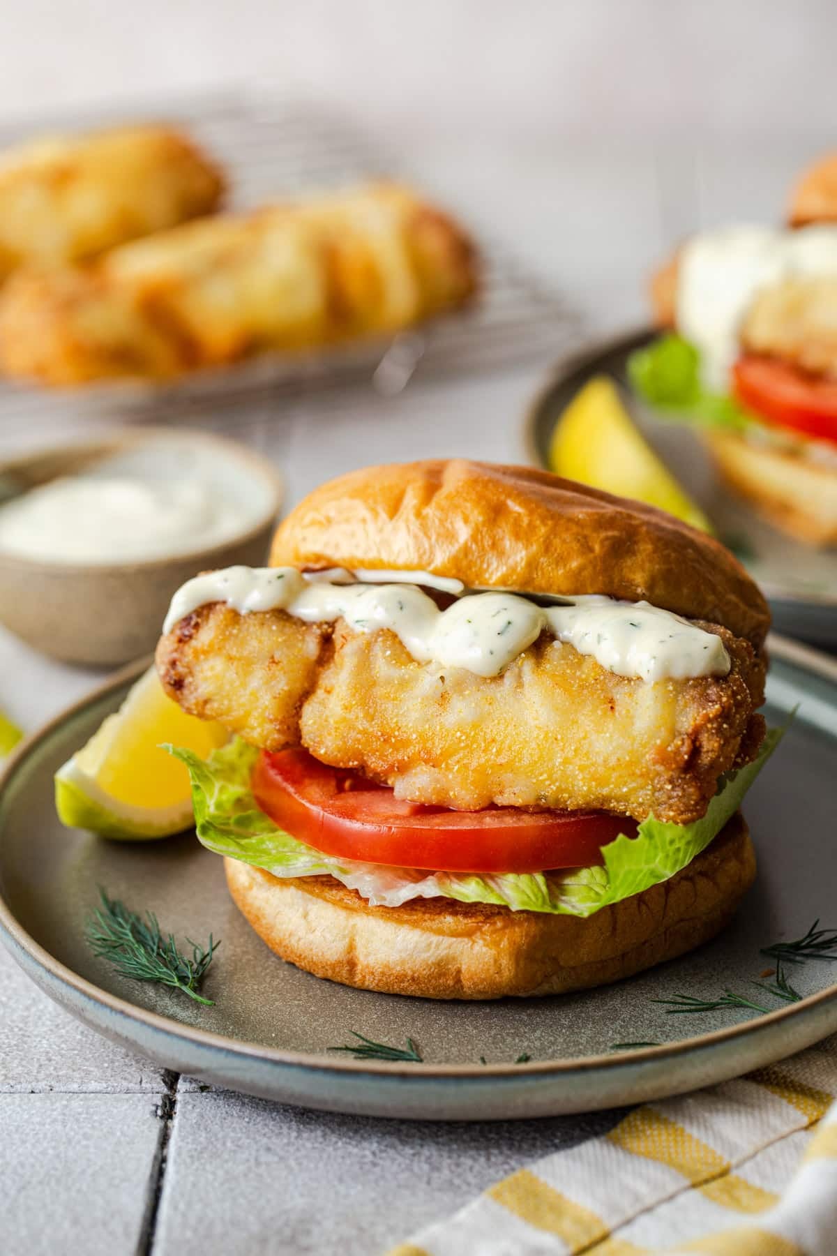 Side shot of a crispy fried fish sandwich on a plate with homemade tartar sauce and a lemon wedge.