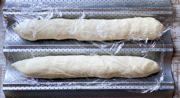 Two loaves of unbaked baguettes sit on an aluminum bread rack, covered in plastic wrap as they proof on a countertop.