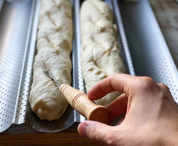 A baker uses a bread lame to cut slices on the surface of two unbaked loaves of French baguettes.
