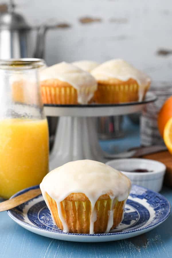 A single orange muffin topped with a sweet glaze sits on a small blue dessert plate. Behind the plate is a glass of orange juice and a platter of freshly baked muffins.