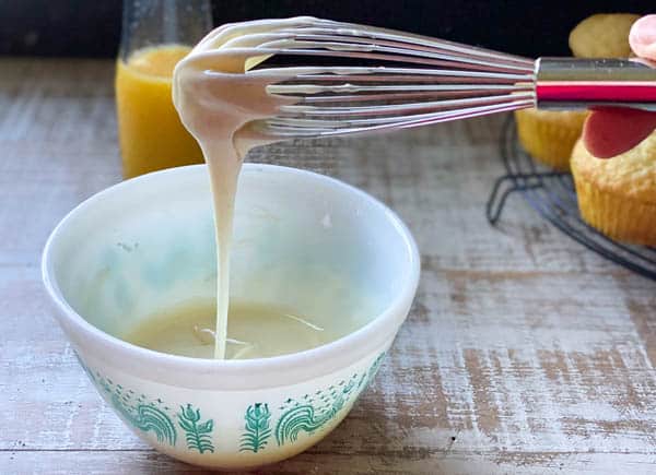 A person holds a metal whisk over a bowl of sweet orange glaze. The glaze drips from the tines of the whisk.