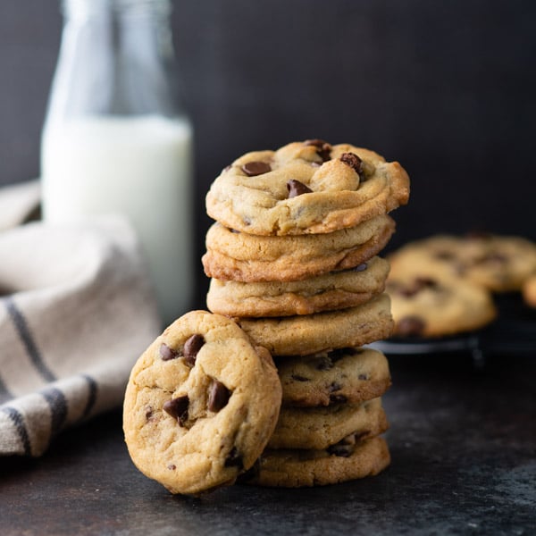 Square image of a stack of homemade chocolate chip cookies with milk in background