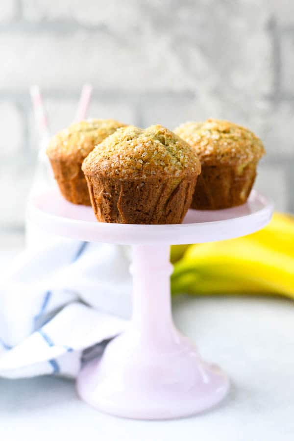 Three freshly baked banana muffins sit on a small light pink cake stand.
