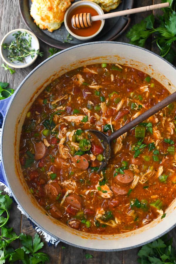 An overhead shot of a Dutch oven filled with freshly cooked chicken and sausage gumbo. The ingredients - shredded chicken, sausage, okra, and other vegetables, and mixed together in a thick tomato-based broth. A wooden serving spoon rests inside the pot of gumbo.