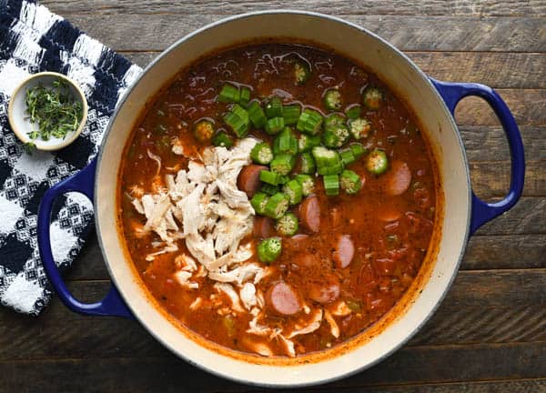 An overhead image of a large Dutch oven, filled with ingredients to make gumbo. Shredded chicken, chopped okra, and sausage cook in a thick tomato-based broth.