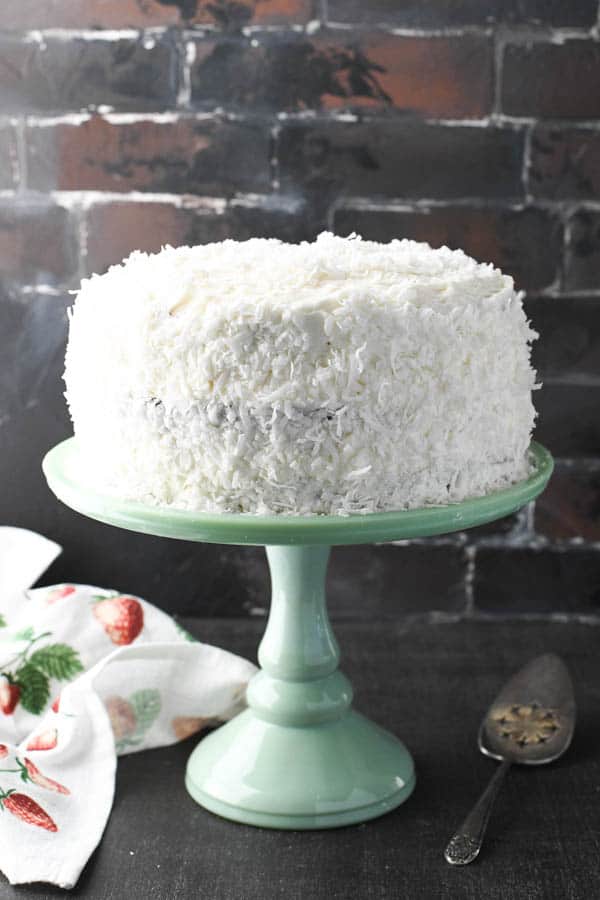 Front shot of a beautiful old fashioned coconut cake on a green cake stand in front of a brick wall