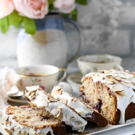 Side shot of a cherry almond loaf cake sliced on a white tray with flowers in the background.