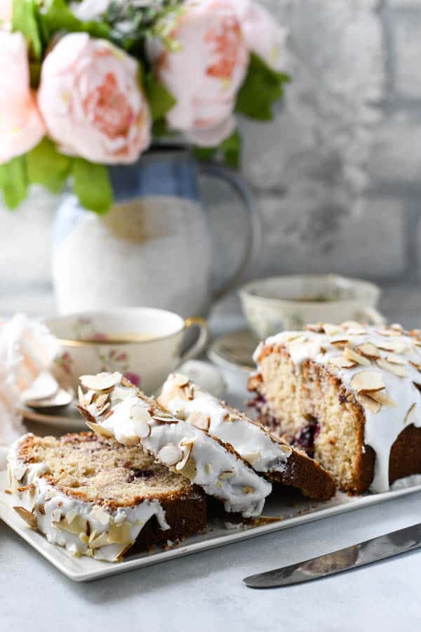 Side shot of a cherry almond loaf cake sliced on a white tray with flowers in the background.