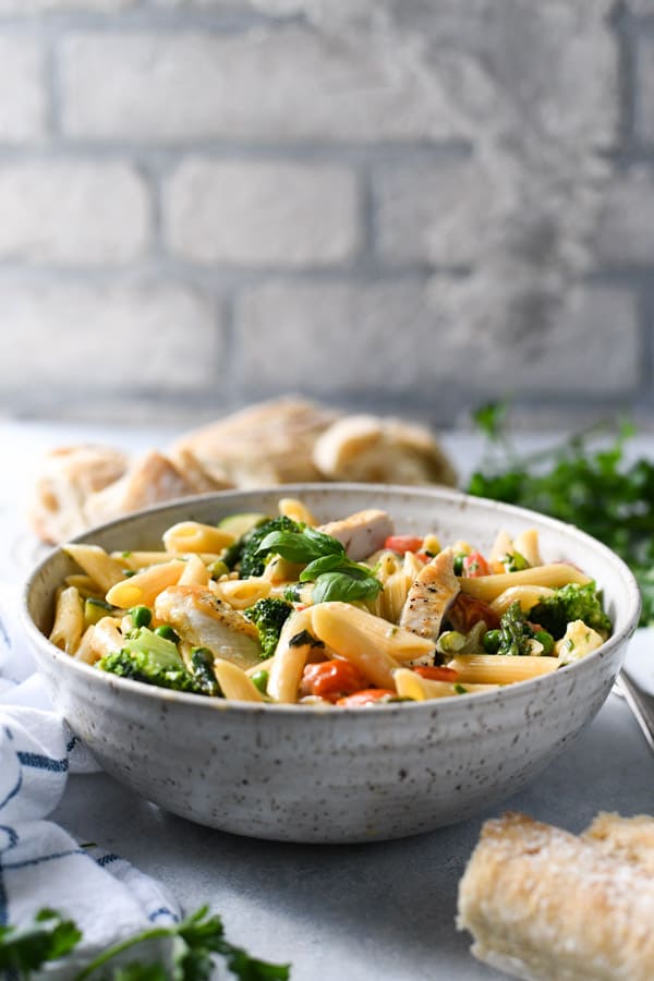 Front shot of a bowl of chicken pasta primavera on a white table in front of a white brick wall