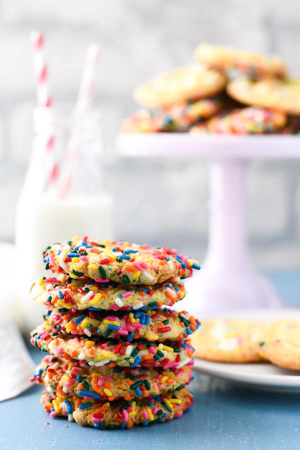 A stack of rainbow sprinkle-covered cookies sit on a blue countertop. Just out of focus in the background is a plate with more cookies and a glass of milk.
