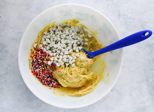 Funfetti cake mix cookie dough mixed together in a white bowl with a blue spatula. Funfetti morsels and sprinkles sit on top of the dough, unmixed.