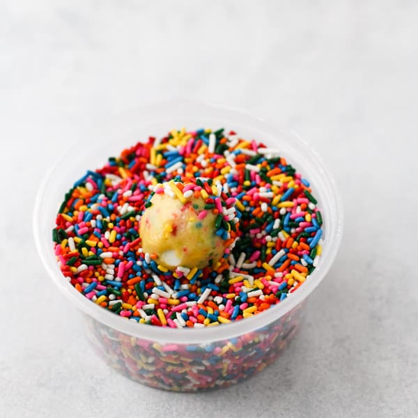 A ball of funfetti cookie dough sits in a container of rainbow sprinkles, covering half of the ball of dough.