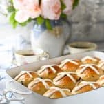 Side shot of the softest hot cross buns recipe on a white table in front of a vase of flowers