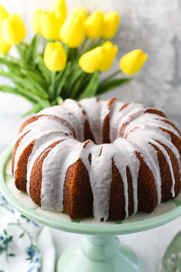 Lemon pudding bundt cake on a green cake stand in front of a vase of yellow tulips