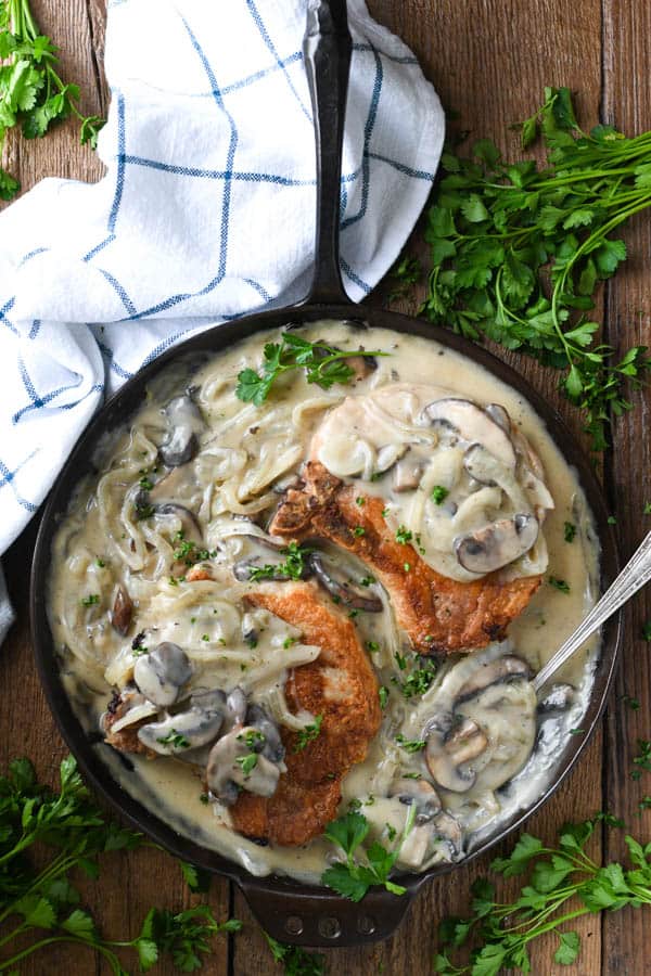 Overhead image of smothered pork chops and gravy in a cast iron skillet on a wooden table with parsley garnish