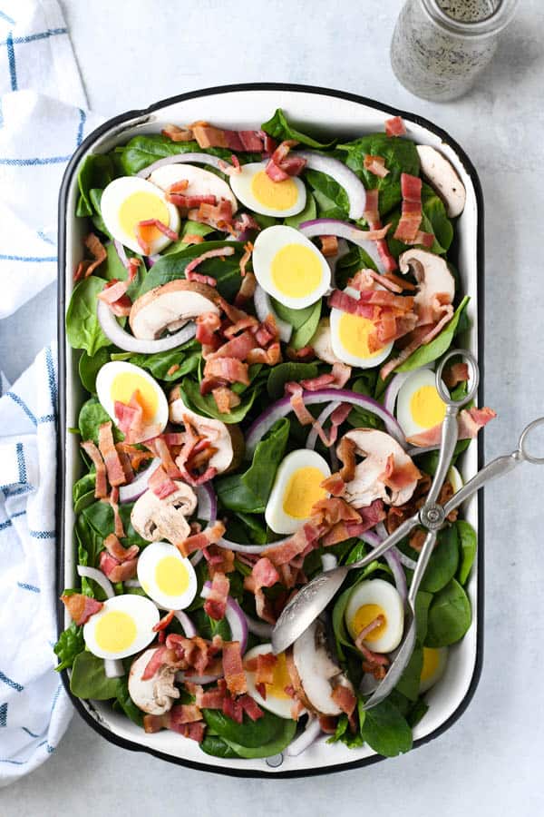 Overhead shot of a spinach salad recipe served in a large dish with eggs, bacon mushrooms and onion