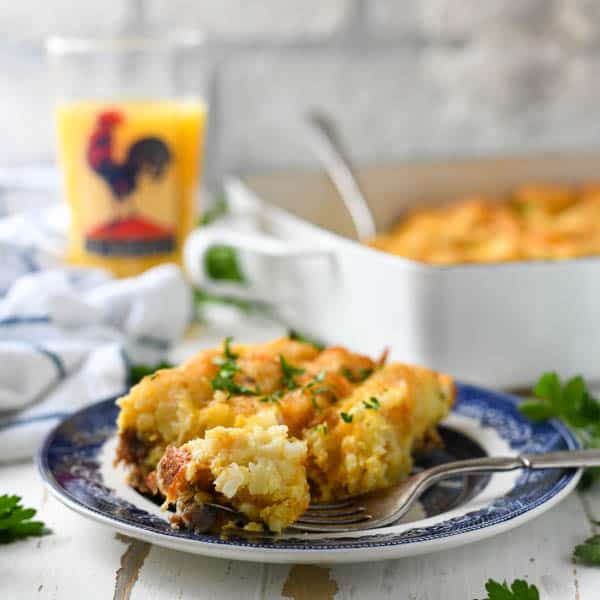 Square image of a slice of tater tot breakfast casserole with sausage on a plate with orange juice in the background