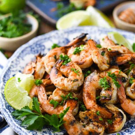 Close up side shot of the best grilled shrimp recipe garnished with parsley and limes on a blue and white plate