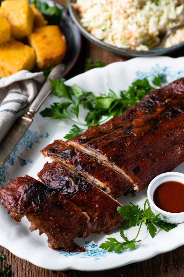 A tray of oven baked baby back ribs coated in barbecue sauce and served with cornbread and coleslaw.
