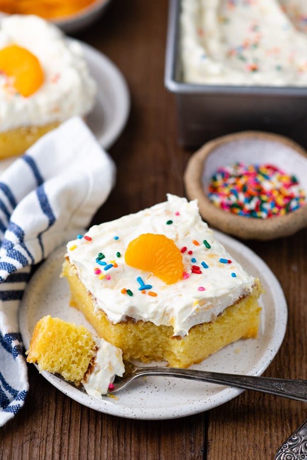 A single slice of Pig Pickin' Cake, garnished with a mandarin orange slice and colorful sprinkles. A silver fork rests on the small plate holding a bite of cake.