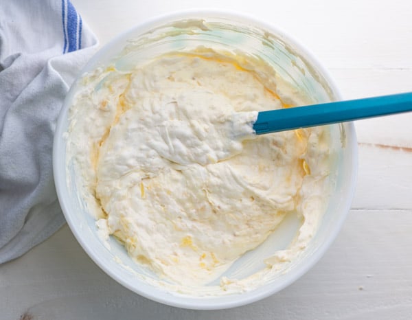 An overhead image of the fluffy pineapple whipped topping for the Pig Pickin' Cake in a white bowl, mixed with a blue spatula.