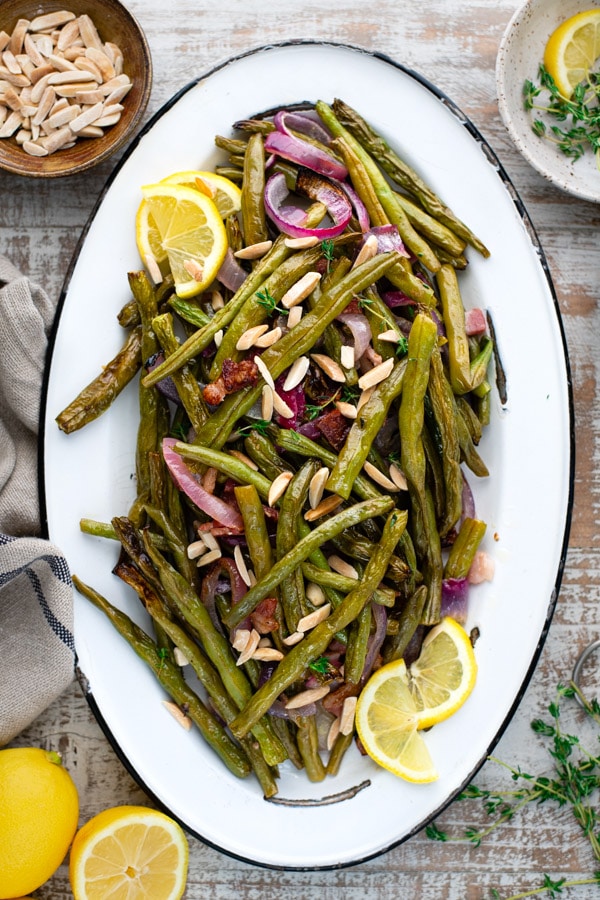Overhead platter of roasted green beans with bacon and herbs on a white surface