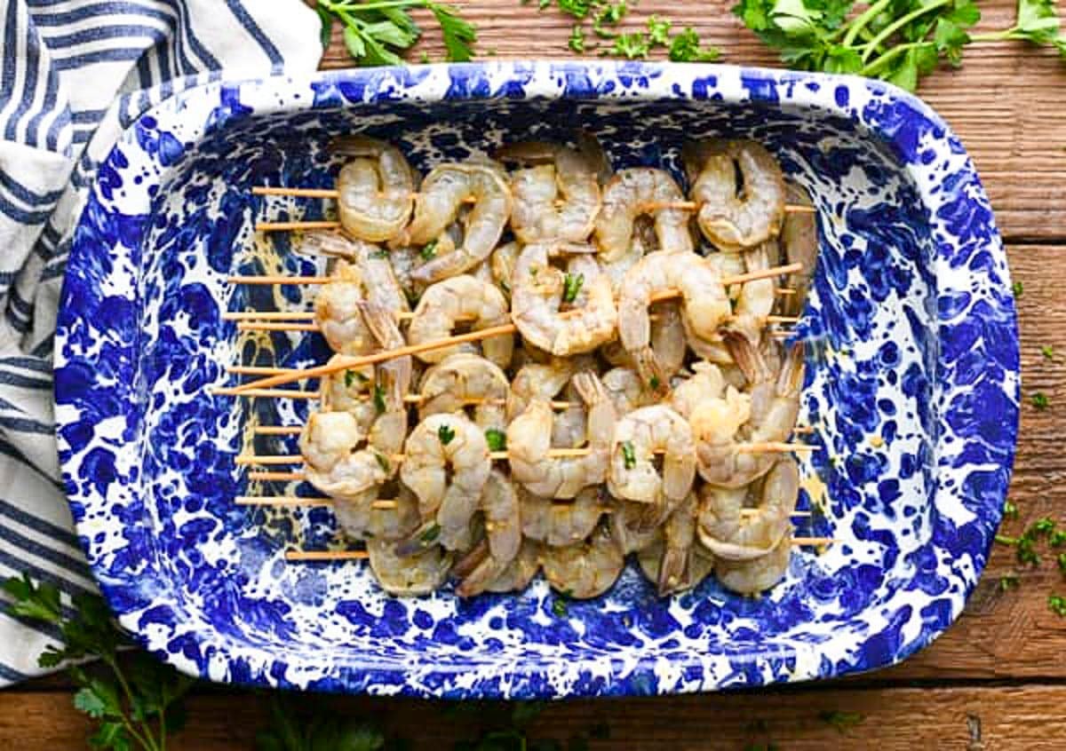 Overhead shot of marinated shrimp on wooden skewers in a blue and white dish before grilling.