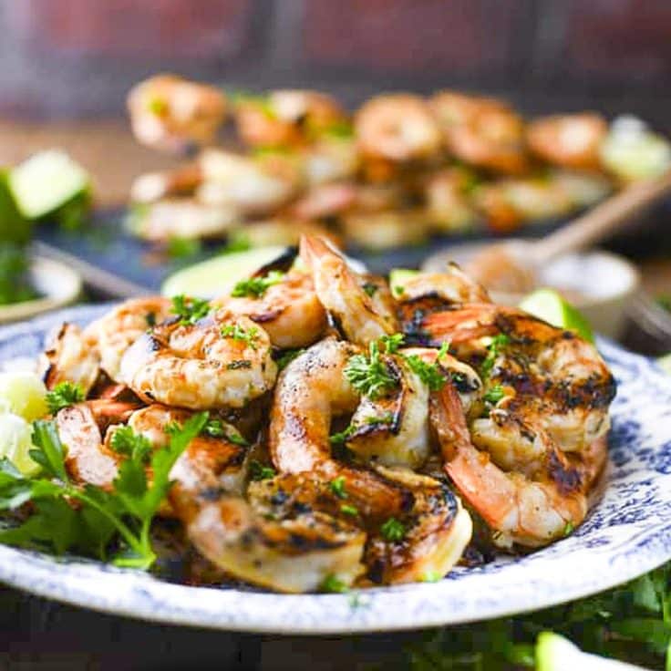 Square side shot of marinated grilled shrimp on a blue and white plate with fresh parsley and lime wedges for garnish.