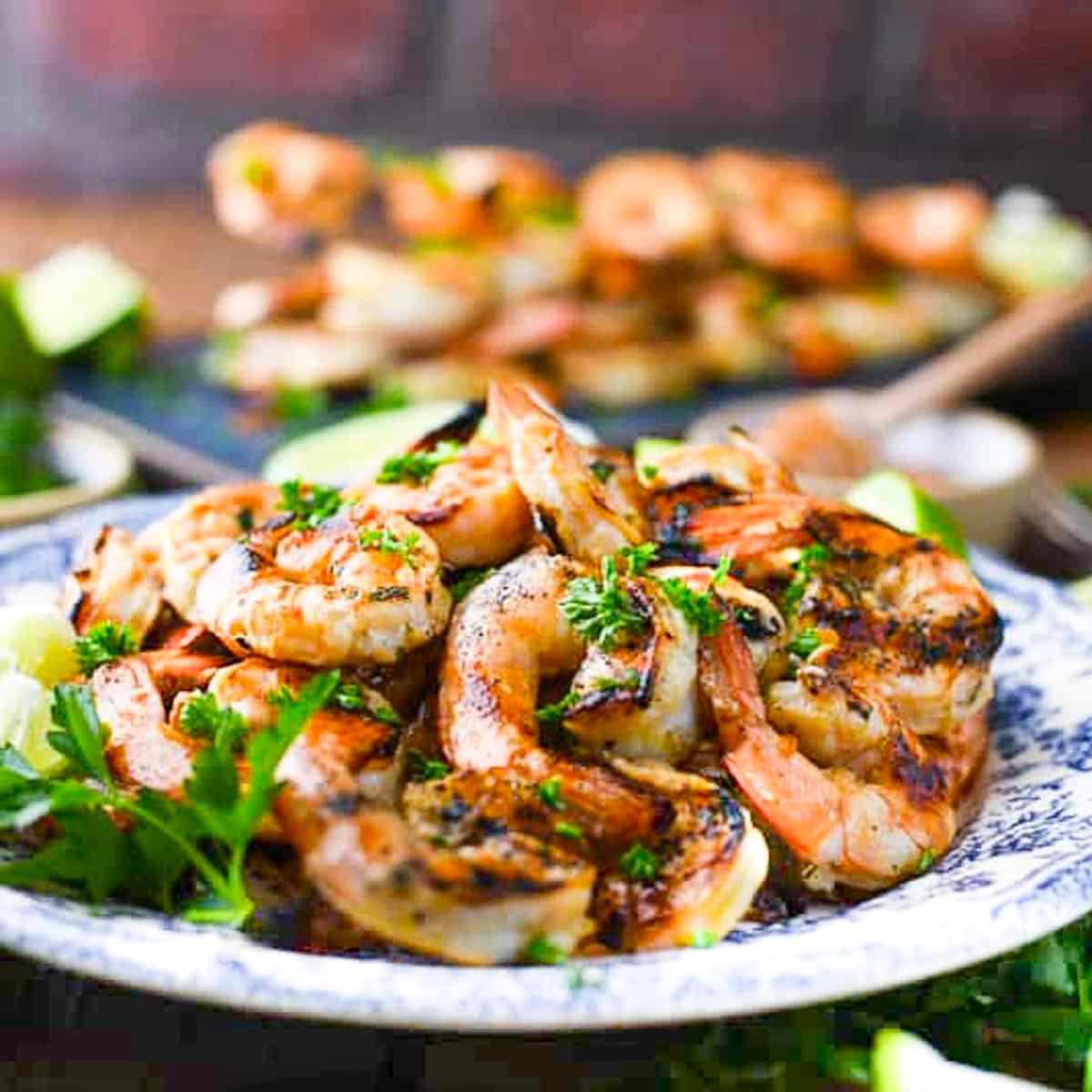 Square side shot of marinated grilled shrimp on a blue and white plate with fresh parsley and lime wedges for garnish.