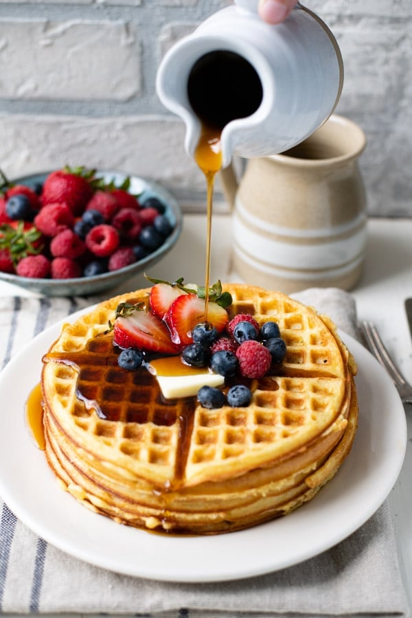 Pouring syrup on a plate of crispy Bisquick waffles with fresh berries