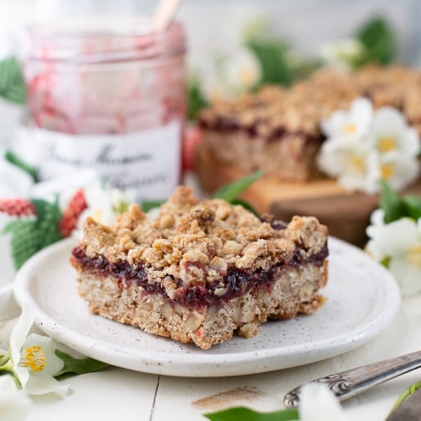 Square image of buttery raspberry crumble bars on a white table with a jar of jam in the background