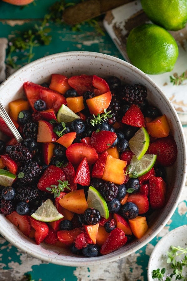 An overhead image of a bowl filled with fresh fruit salad, made with sliced strawberries, blueberries, blackberries, peaches, and line slices.