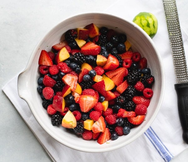 An overhead shot of mixed fruit in a bowl. The fruits include strawberries, raspberries, blueberries, blackberries, and peaches. A zester sits on the countertop next to a zested lime.