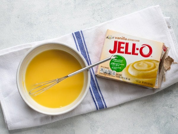 A box of vanilla instant Jello pudding mix sits next to a small bowl filled with a fruit salad dressing. The dressing is made by combining vanilla pudding mix with lime juice and mixing with a small metal whisk.