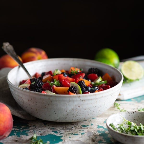 A side shot of a white ceramic bowl filled with a fresh fruit salad. Whole peaches and limes sit on the countertop surrounding the bowl.