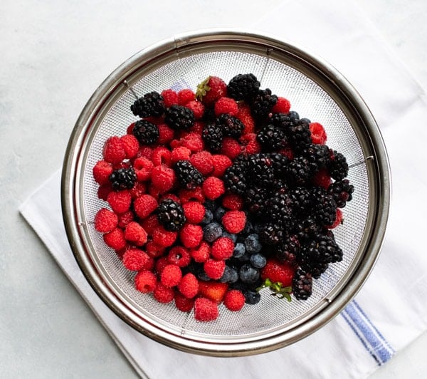 A mixture of fresh raspberries, blueberries, and blackberries sit in a wire strainer over a white paper towel.