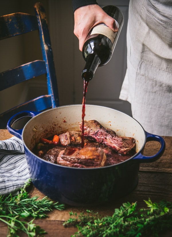 A woman pours a bottle of red wine into a dutch oven filled with beef short ribs and chopped vegetables. Springs of fresh herbs sit on a wooden table surrounding the stock pot.