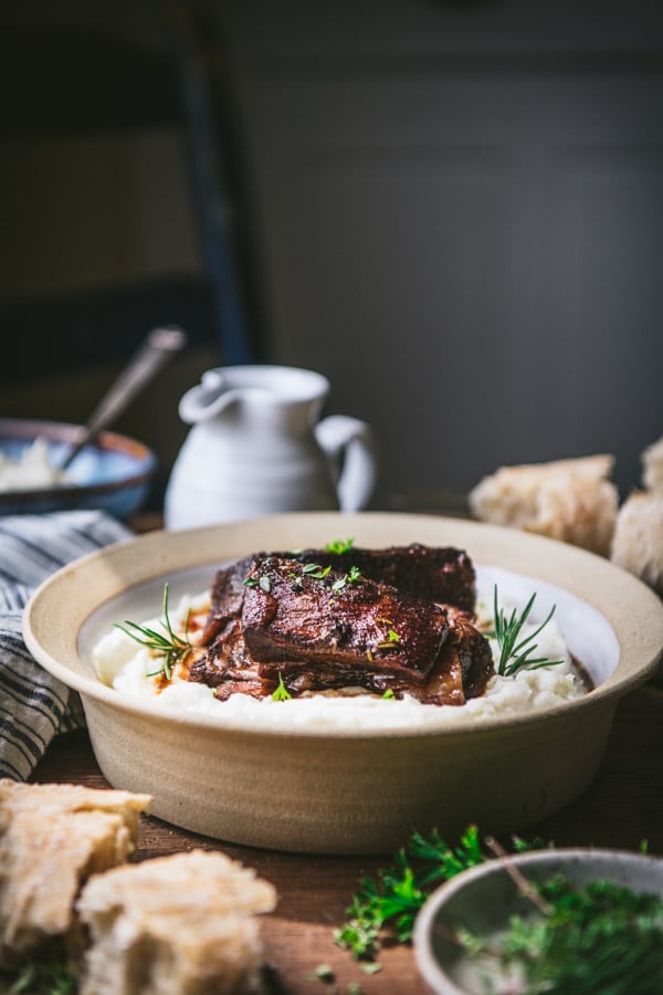 A bowl of red white braised beef short ribs served with mashed potatoes. The bowl sits on a wood table surrounded by pieces of baguette, fresh herbs, and dishes.