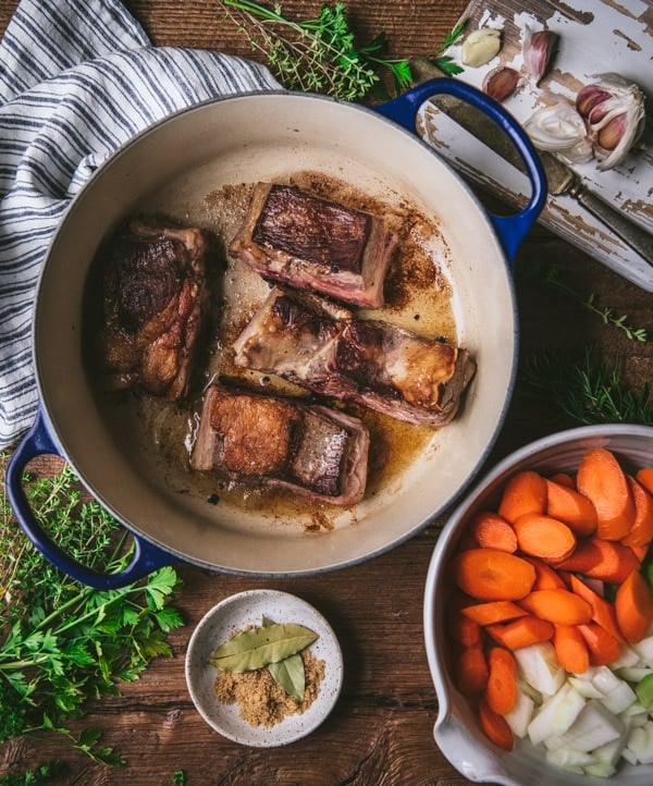 A blue cast iron dutch oven filled with beef short ribs. In this overhead image, there's also fresh herbs and seasonings and a bowl of chopped onions and carrots.
