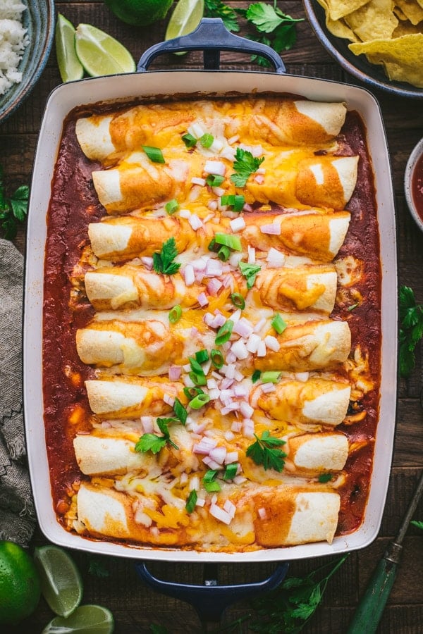 A casserole dish of homemade easy chicken enchiladas baked in a zesty red sauce and topped with melted cheese, green and red onions, and fresh cilantro.