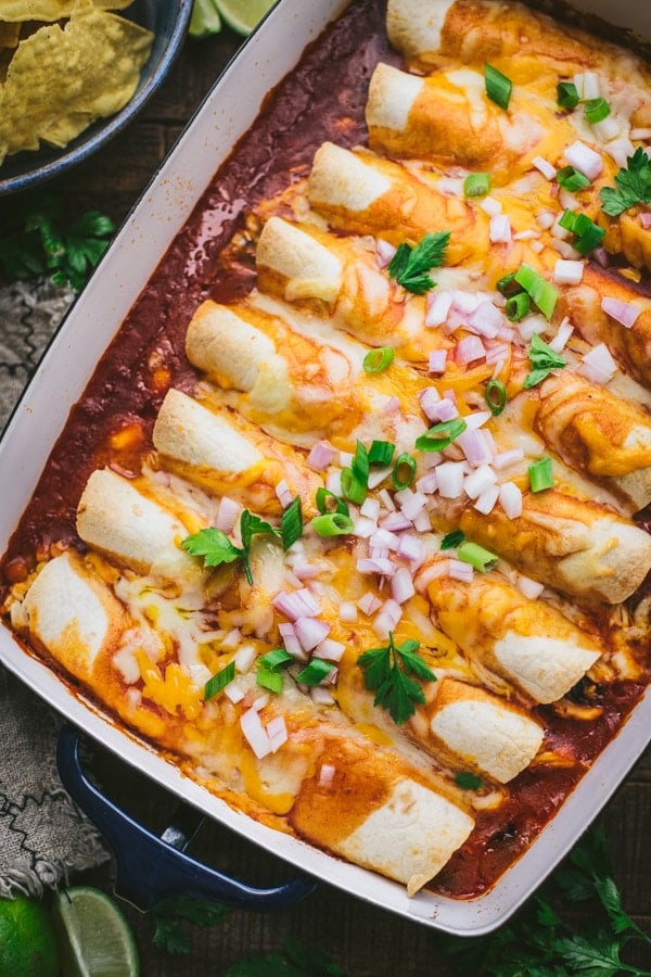 An overhead image of a row of rolled chicken enchiladas in a casserole dish, baked in red enchilada sauce and topped with cheese, onions, and fresh cilantro.