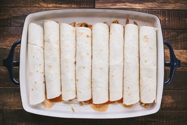 Neatly rolled flour tortillas filled with chicken enchilada mix, lined up in a casserole dish on a wooden cutting board.