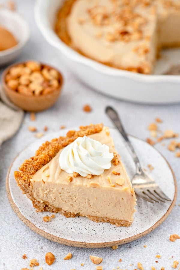Big slice of the best peanut butter pie recipe served on a small white plate with a dollop of whipped cream on top. Chucks of crushed up peanuts are scattered around the plate and on top of the pie.