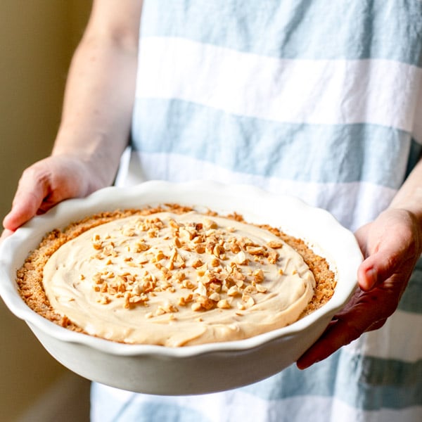 A woman is holding a no-bake peanut butter pie, topped with chopped peanuts in a white scalloped-edge pie dish.