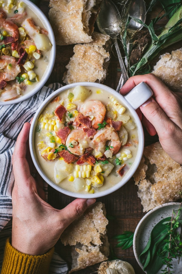 Overhead shot of hands holding a bowl of creamy shrimp soup with corn and bacon