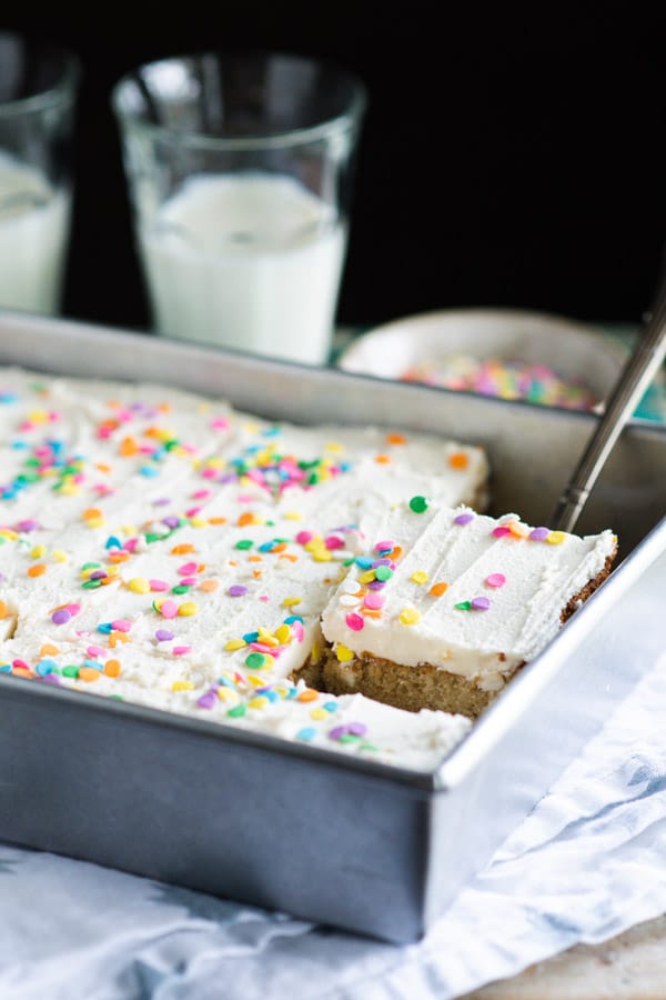 Cutting a single slice from a sheet pan filled with homemade vanilla cake, topped with vanilla buttercream frosting and rainbow confetti sprinkles.