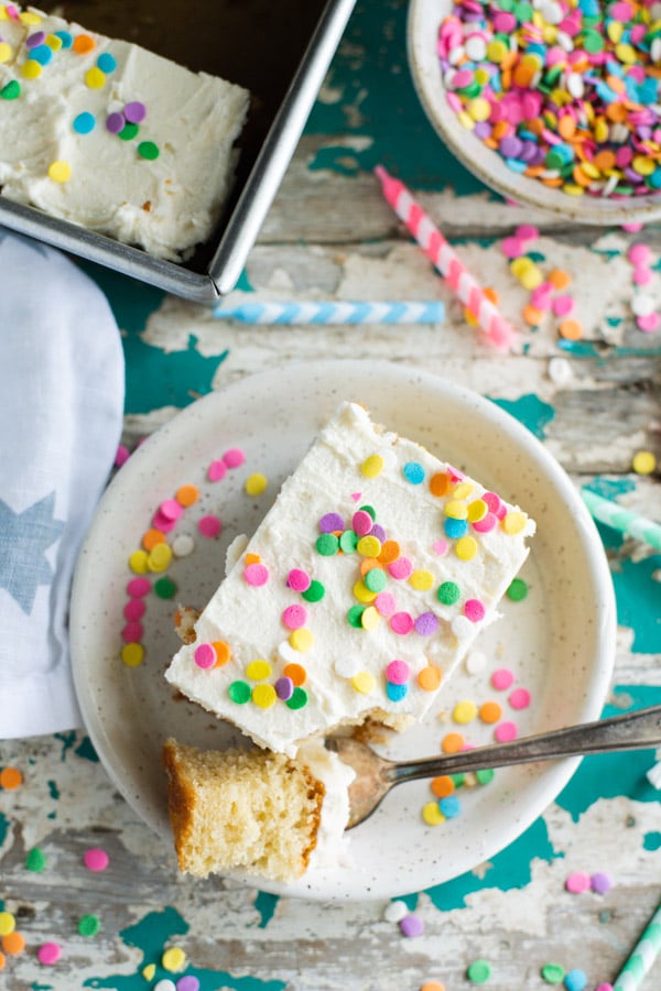 An overhead shot of a single piece of homemade vanilla cake on a plate, topped with vanilla buttercream and colorful rainbow confetti sprinkles. 