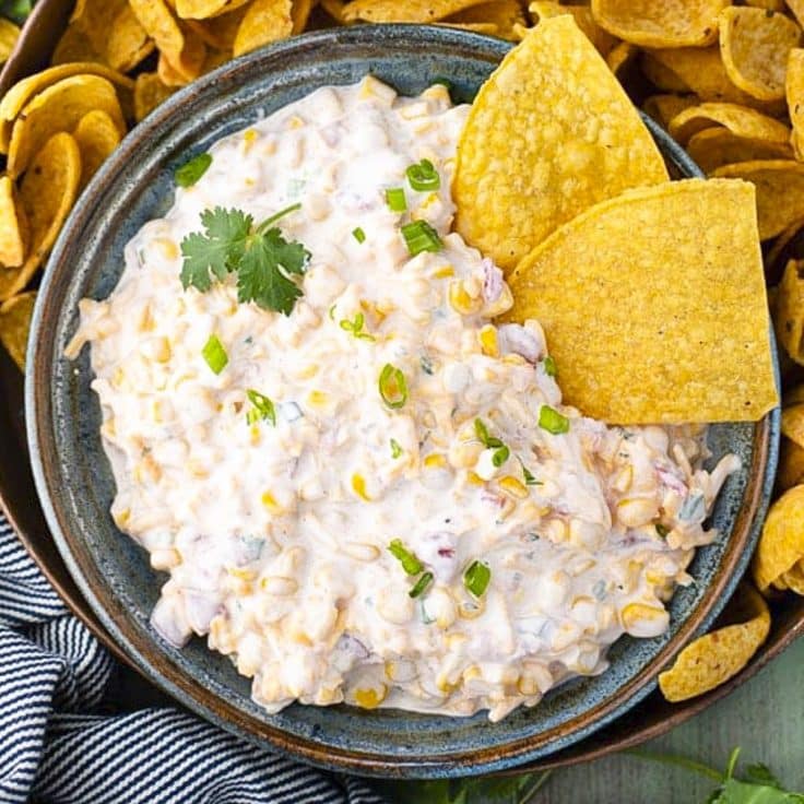A bowl of creamy cold cheesy corn dip with tortilla chips for scooping.